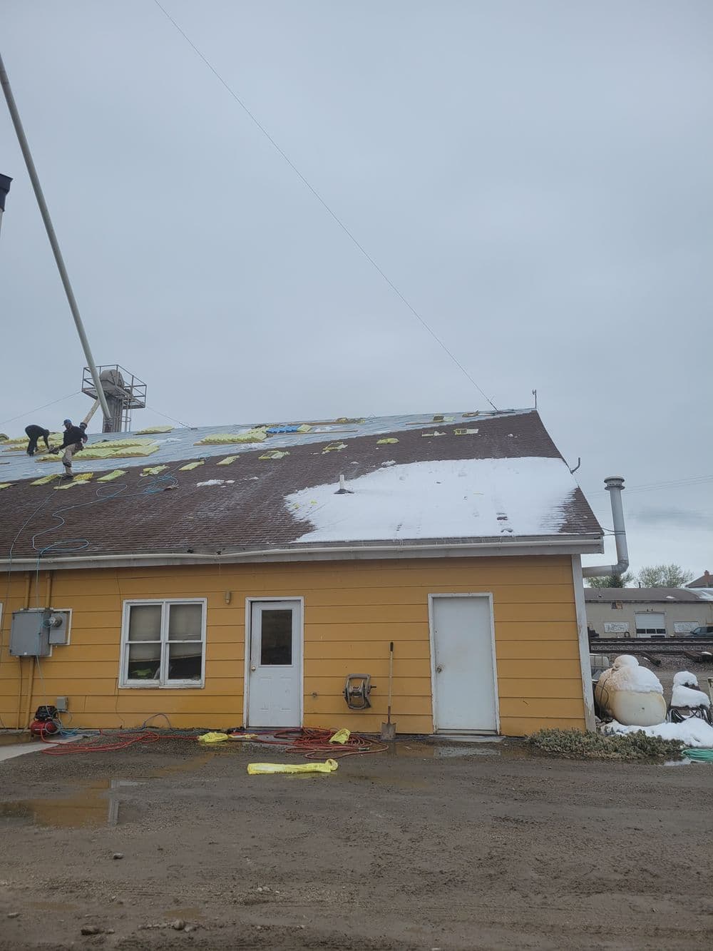 Workers applying new shingles to a roof with snow, yellow tarps, and equipment nearby.