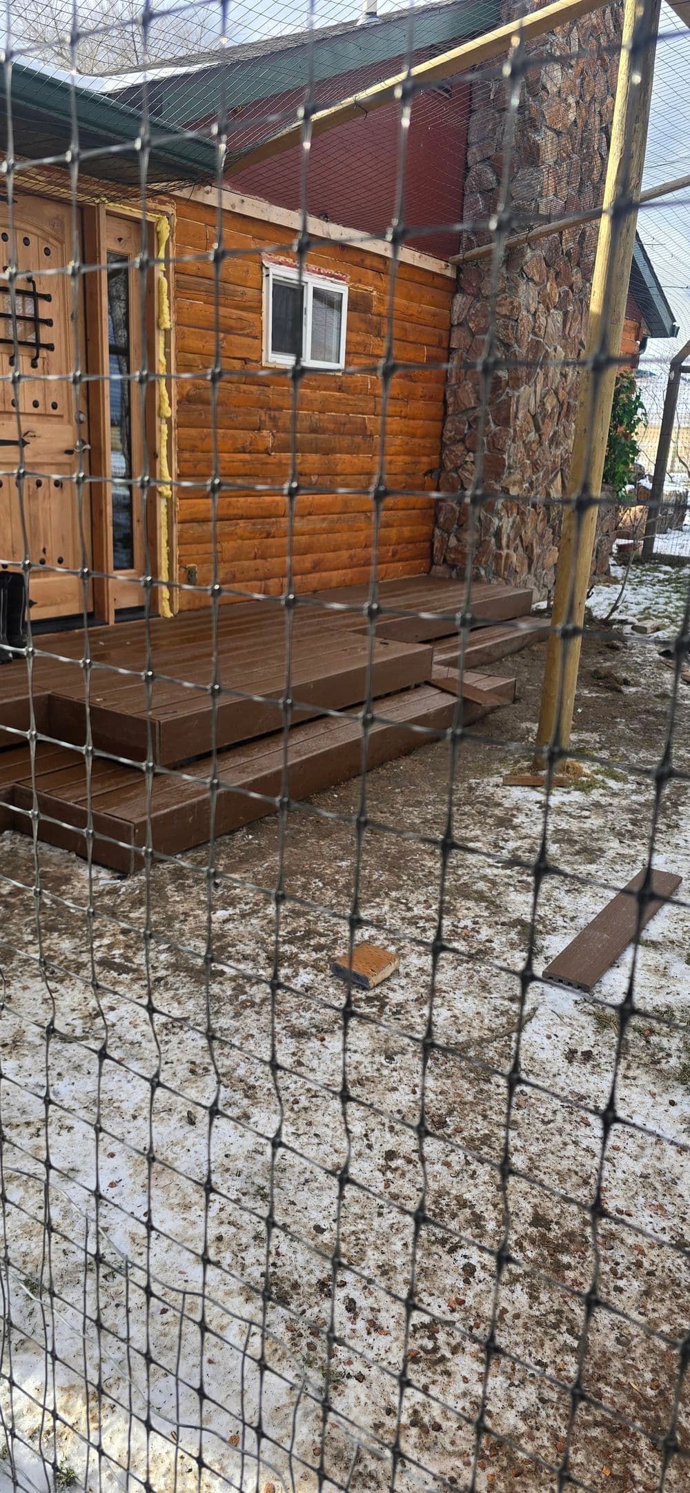 Wooden deck leading to a log cabin, surrounded by a wire fence and snowy ground.
