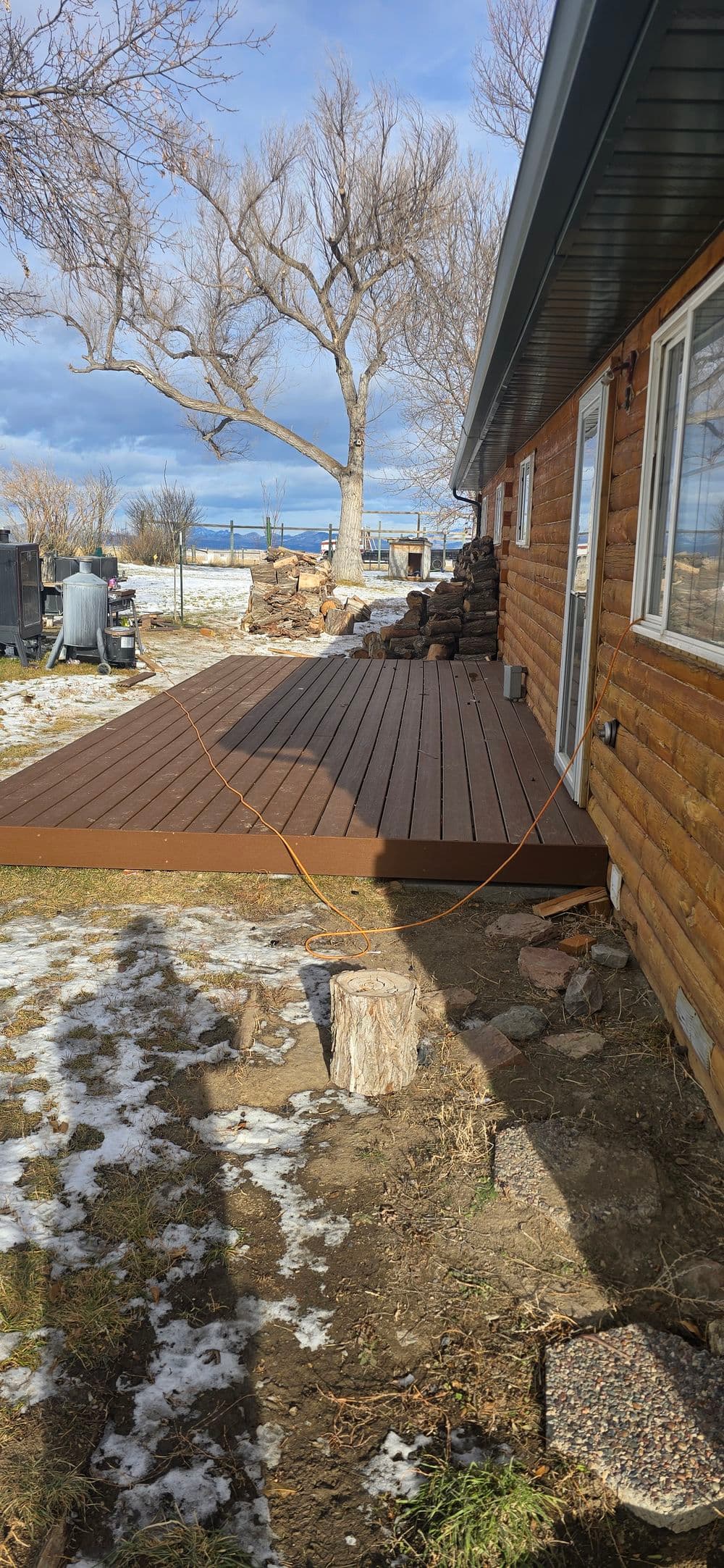Wooden deck extension beside log cabin, with snowy ground and clear blue sky.