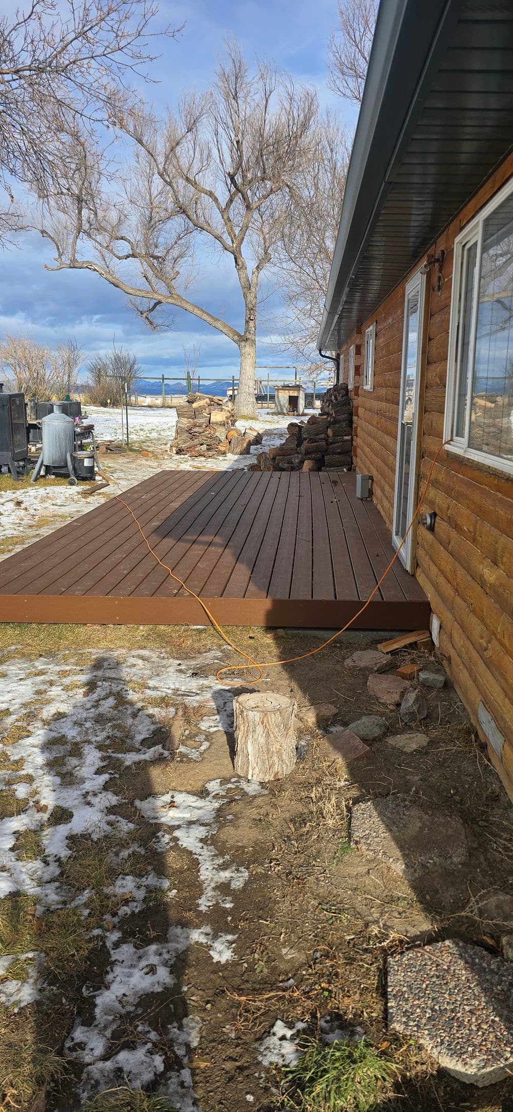 Wooden deck next to a log cabin, snowy ground, and distant trees under a cloudy sky.