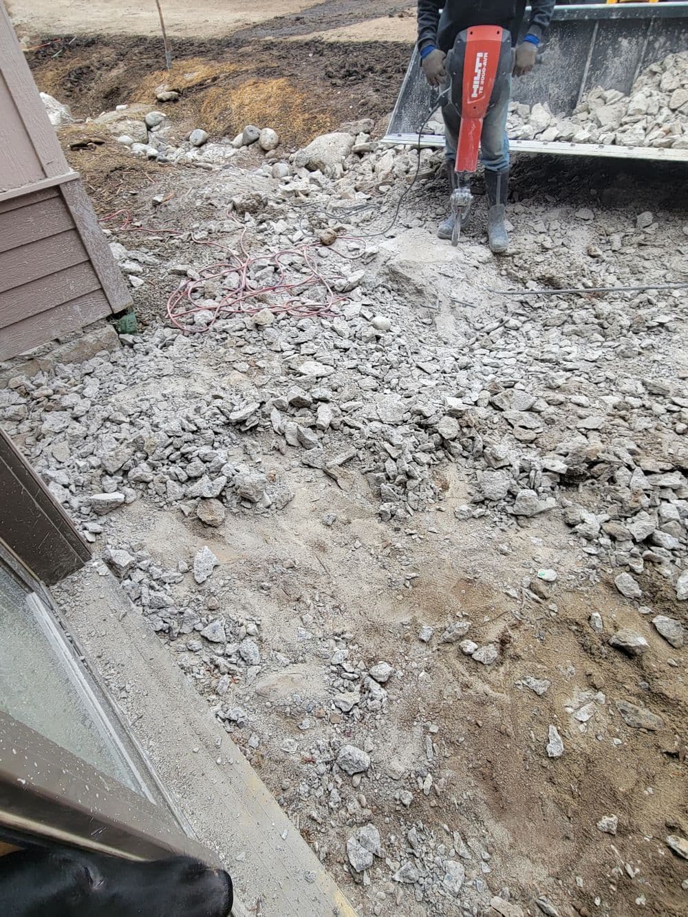 Construction worker using a jackhammer on a gravel site near a building.