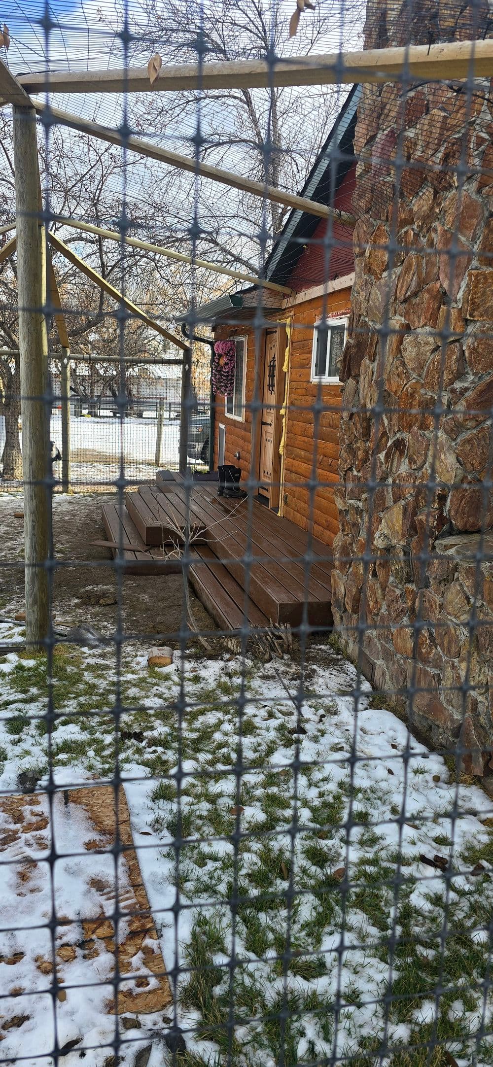 Log cabin exterior with stone wall, wooden deck, and snow-covered ground, framed by wire fence.