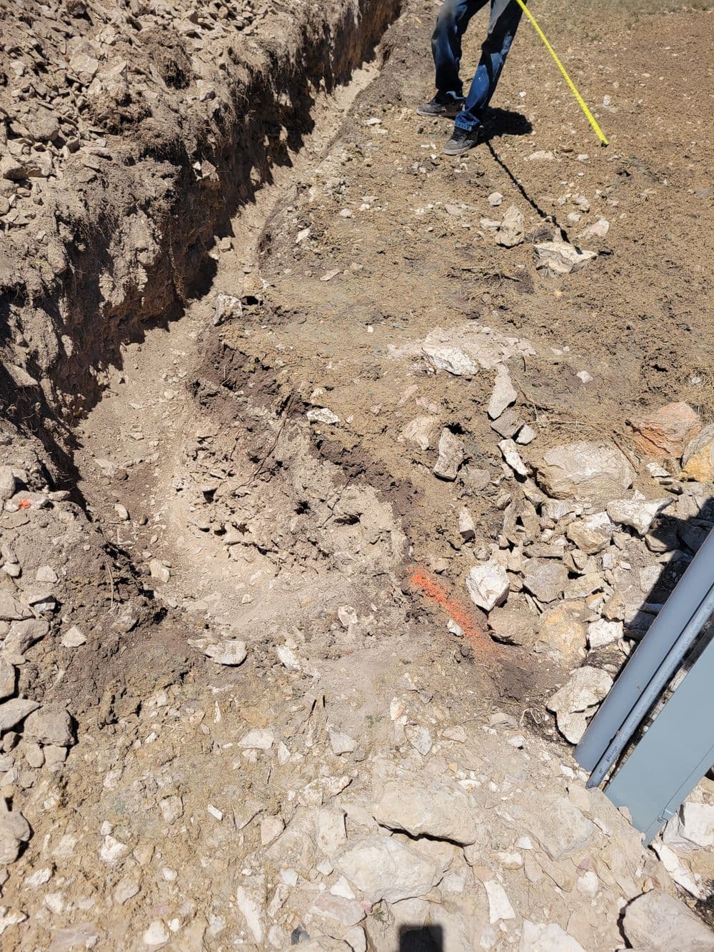 Excavation site showing rocky soil and measurement being taken with a tape measure.