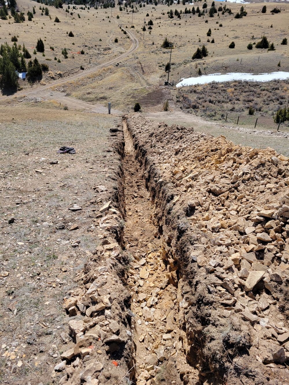 Excavated trench with rocky soil in an open landscape near a rural road.