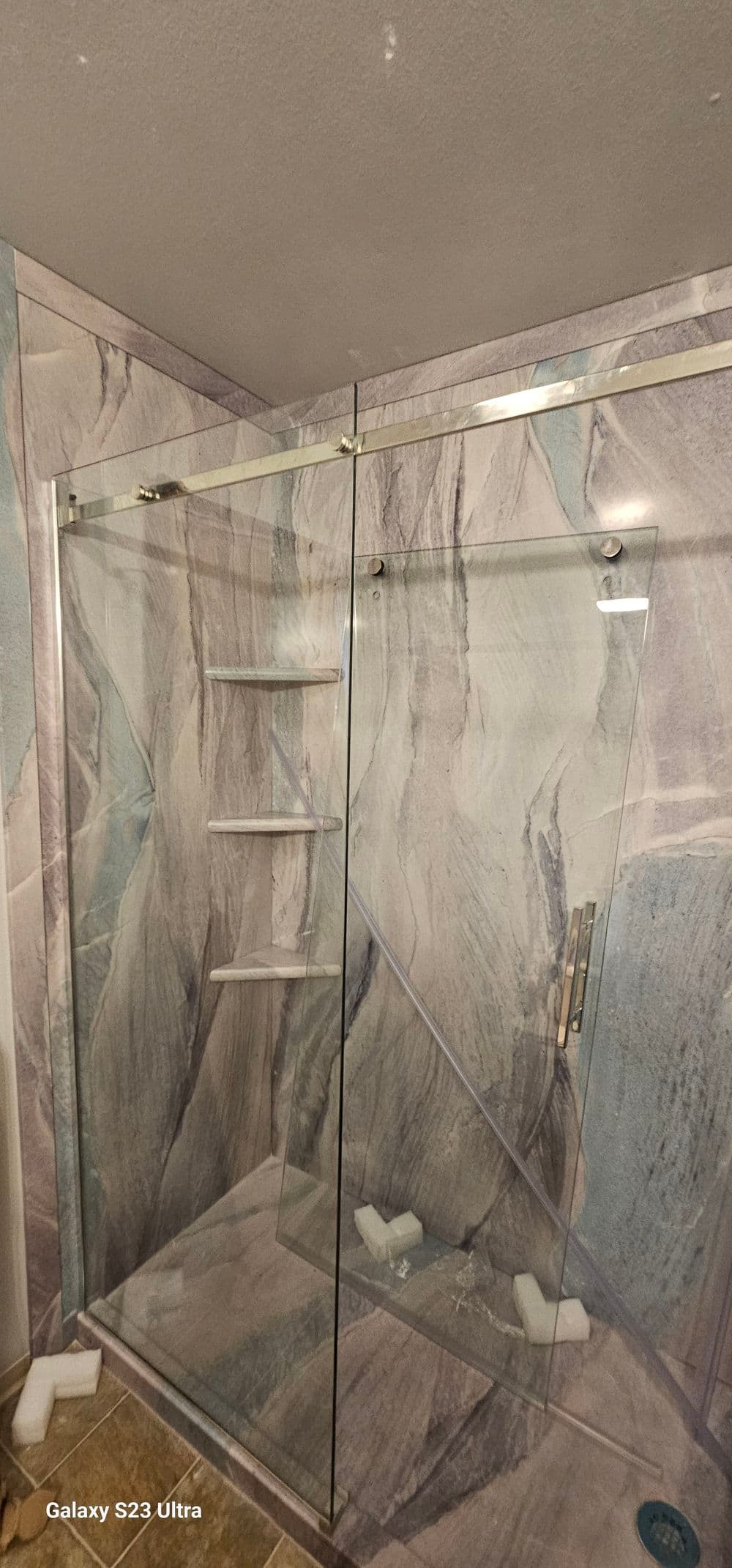 Modern glass shower enclosure with marble-patterned wall and shelves inside a bathroom.